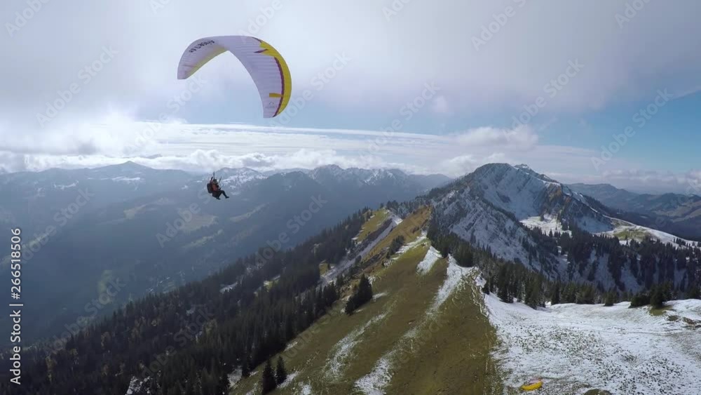 Drone shot of a man paragliding above a mountain ridge, with other mountains and forests in the background, on a sunny day with some clouds Drone shot of a man paragliding above a mountain ridge, with other mountains and forests in the background, on a sunny day with some clouds
