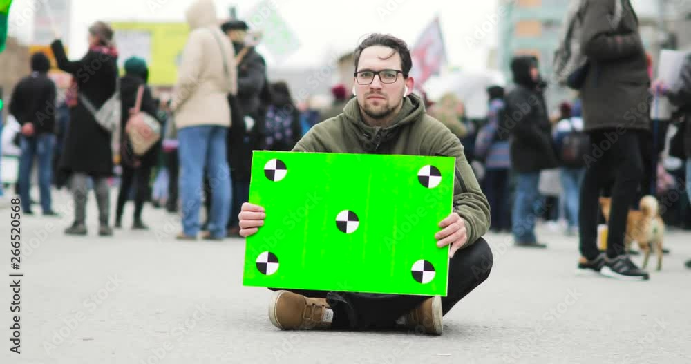 Poster with green screen in man's hands who is sitting on the street during demonstration in city with crowd on background.