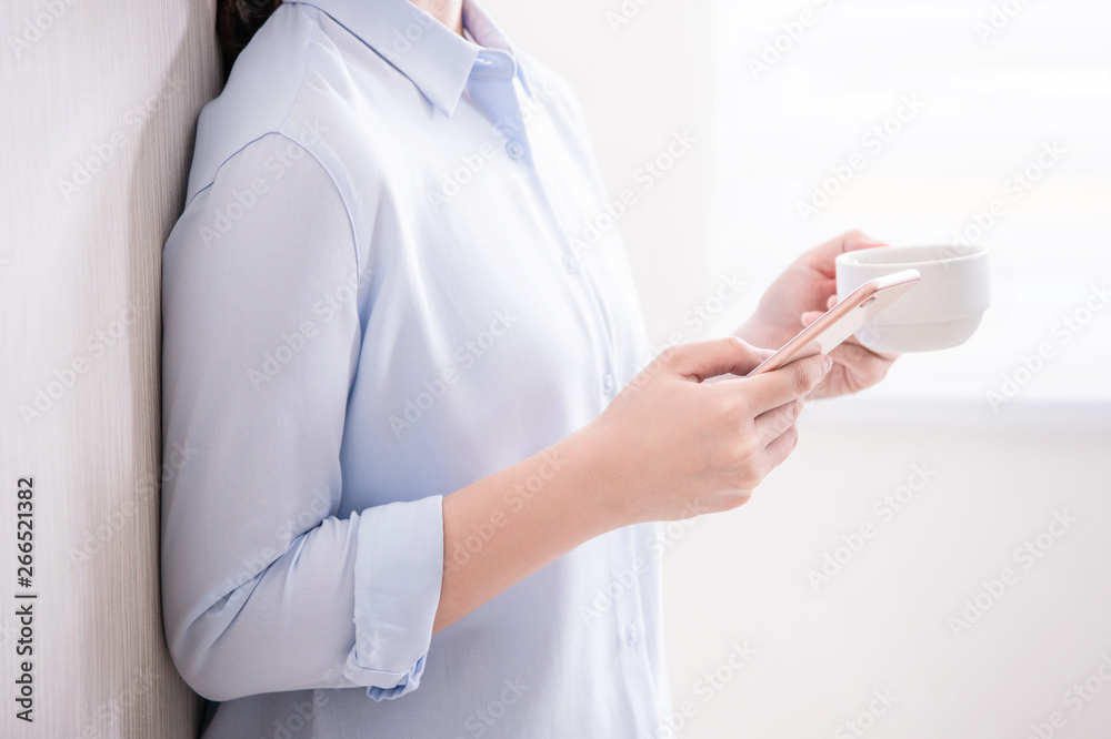 Side view of asia young business woman in blue shirt standing lying on the wall beside window drinking and using smart phone, copy space, close up