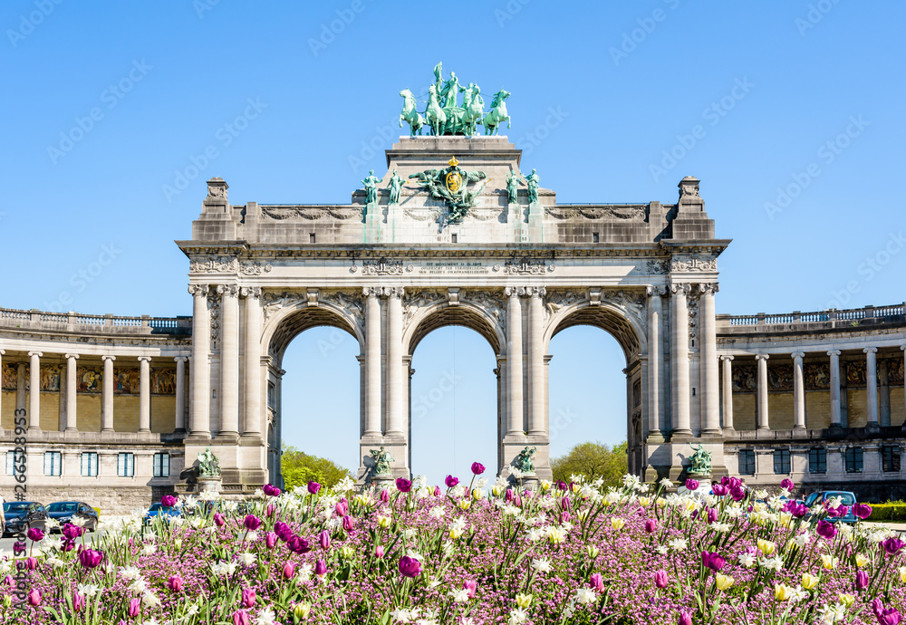 Obraz premium The arcade du Cinquantenaire, the triumphal arch erected by king Leopold II in the Cinquantenaire park in Brussels, Belgium, with a flowerbed in full bloom in the foreground against blue sky.