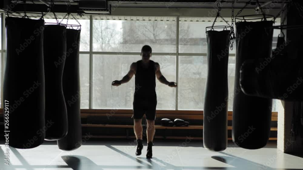 Camera approaching skilled professional boxer doing exercise with jump rope to warm up before practice and punching boxing bag in gym