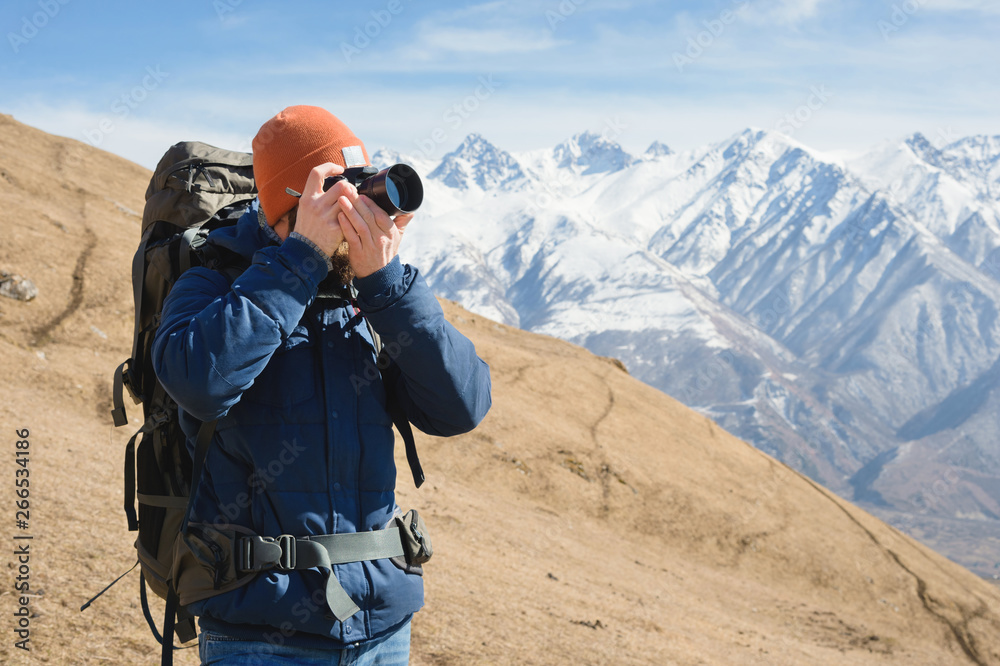 Portrait of a bearded male photographer in sunglasses and a warm jacket with a backpack on his back and a reflex camera in his hands takes pictures against the background of snow-capped mountains