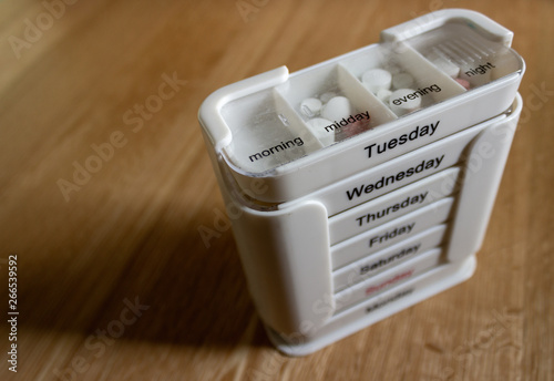 White seven day four compartment medication dispenser with medication standing on an oak surface. Subject lit with natural light from front