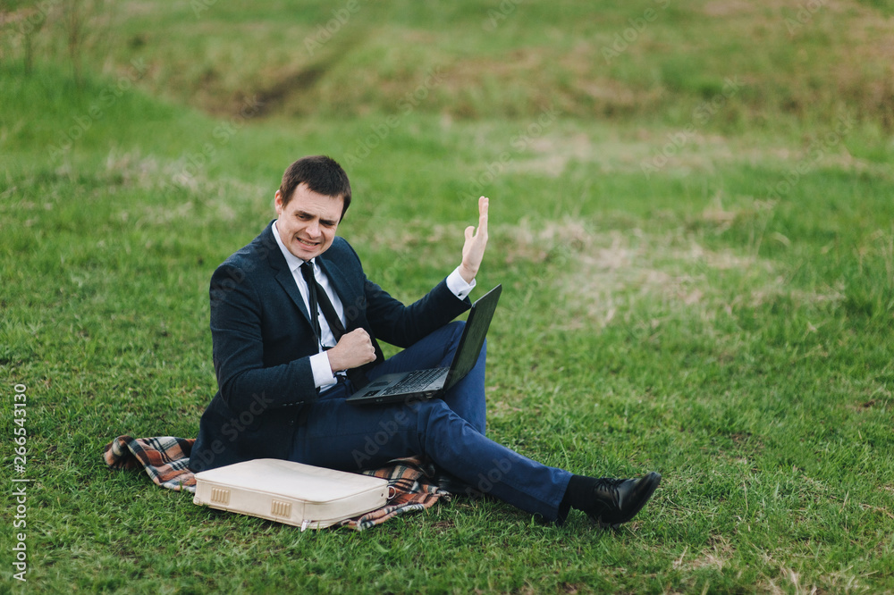 An angry and annoyed businessman in a suit sits in a park in spring, on green grass and works on a laptop. A broken computer, lifestyle, hard work, an IT professional. Negative emotions. An angry and annoyed businessman in a suit sits in a park in spring, on green grass and works on a laptop. A broken computer, lifestyle, hard work, an IT professional. Negative emotions.