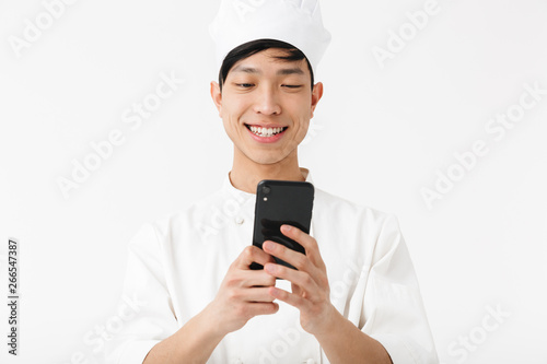 Image of cheerful chinese chief man in white cook uniform and chef's hat holding mobile phone