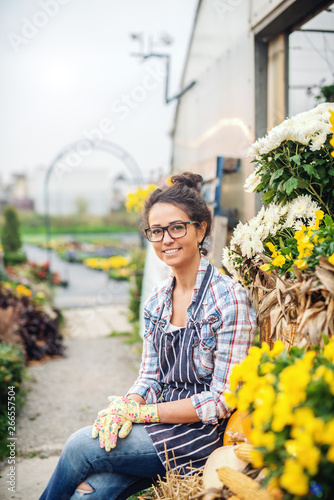 Charming Caucasian florist in apron and gardening gloves on sitting in front of greenhouse and looking at camera.