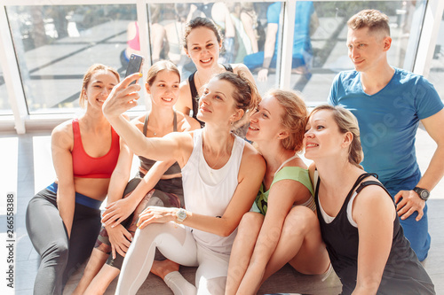 Group of positive young people take a selfie on a smartphone after a group of yoga classes in their bright room with a large window. Concept lovers of yoga and pilates