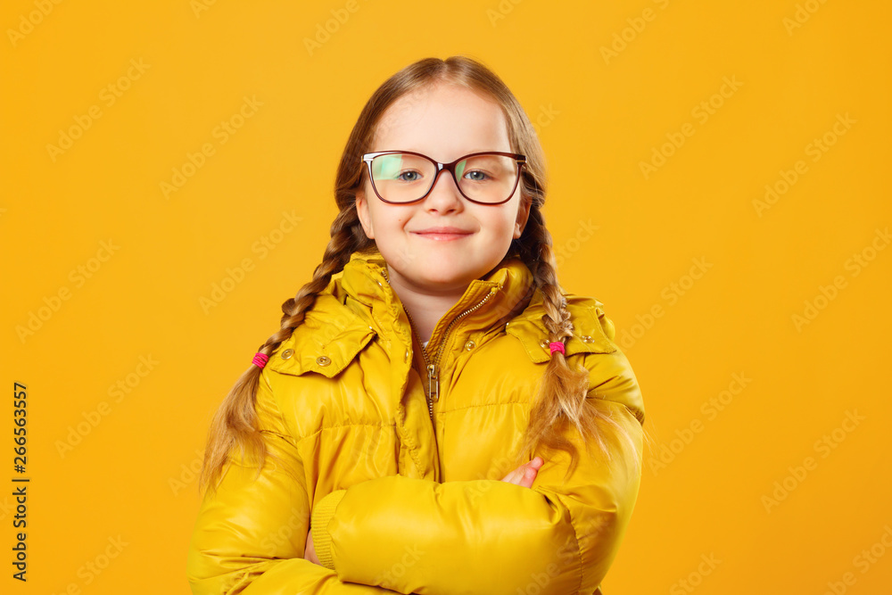 Closeup portrait of a cheerful little girl in jacket and glasses over yellow background. The child folded his arms and looks into the camera.
