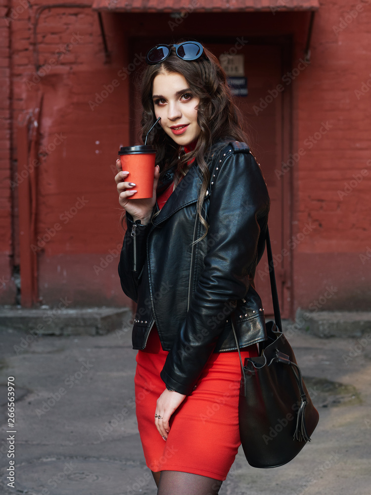 Portrait of a young happy curly brown-haired girl dressed in red dress leather jacket holding coffee cup while standing in of old city. Stock Photo | Adobe