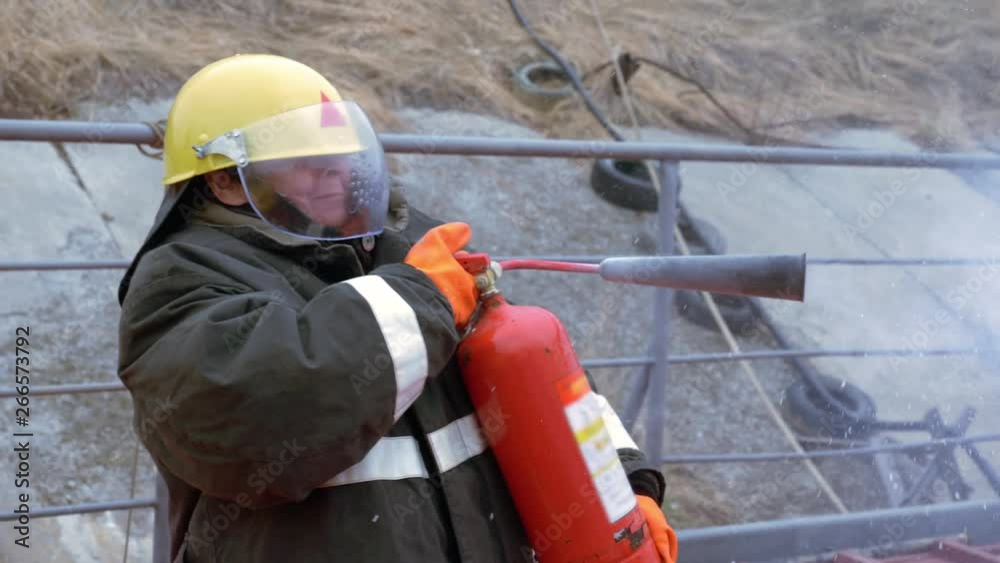 Woman fireman in protective helmet and uniform extinguishes flame with extinguisher in barbeque grill. Fireman with extinguisher fighting fire during training. Female emergency workers Woman fireman in protective helmet and uniform extinguishes flame with extinguisher in barbeque grill. Fireman with extinguisher fighting fire during training. Female emergency workers