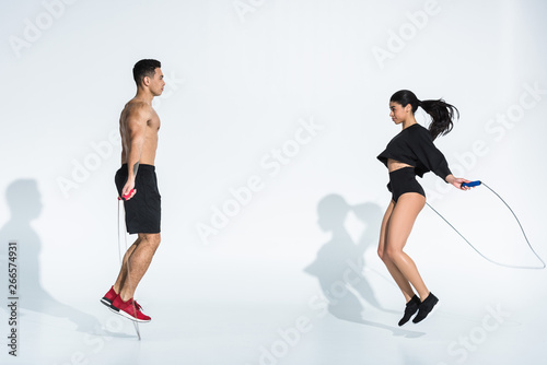 sportive multicultural man and woman jumping with skipping ropes on white
