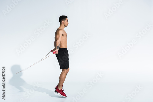 side view of sportive mixed race man in black shorts and red sneakers jumping with skipping rope on white