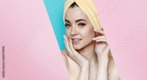 Portrait of young beautiful woman with healthy glow perfect smooth skin look into the hole of pink paper on blue background. Model with natural nude make up with yellow towel on head. © KDdesignphoto