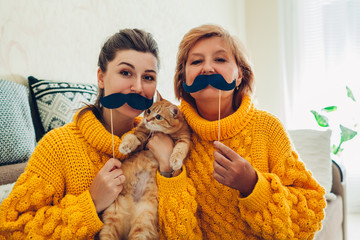 Senior mother and her adult daughter taking selfie with cat using photo booth props at home. Mother's day concept.
