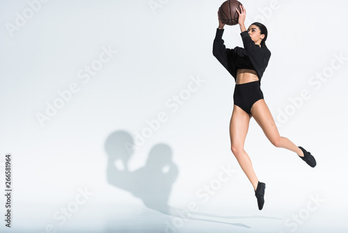 sportive african american girl in black sportswear and sneakers jumping with ball on white background