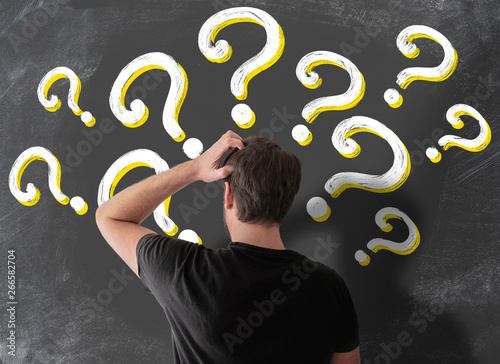 rear view of puzzled man in t-shirt scratching his head in confusion against blackboard filled with question marks photo