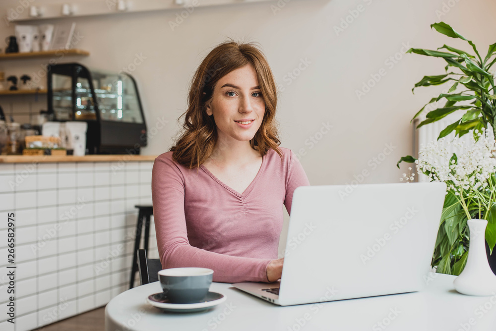 Beautiful young redhead woman working with laptop in cafe and drinking coffee Beautiful young redhead woman working with laptop in cafe and drinking coffee