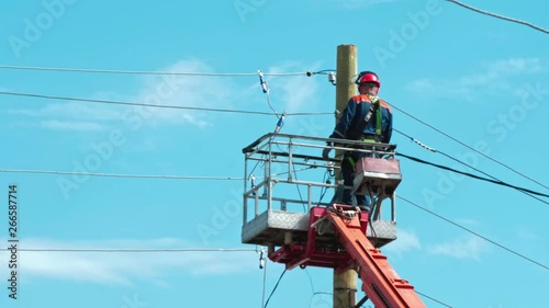 Electrician in uniform repairs power lines, standing on the bucket. Summer day. In the background is a blue cloudy sky.