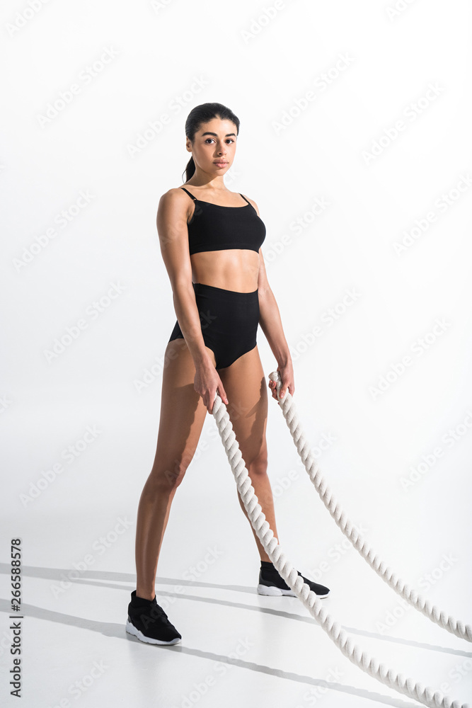 beautiful sportive african american girl holding white ropes and looking at camera on white
