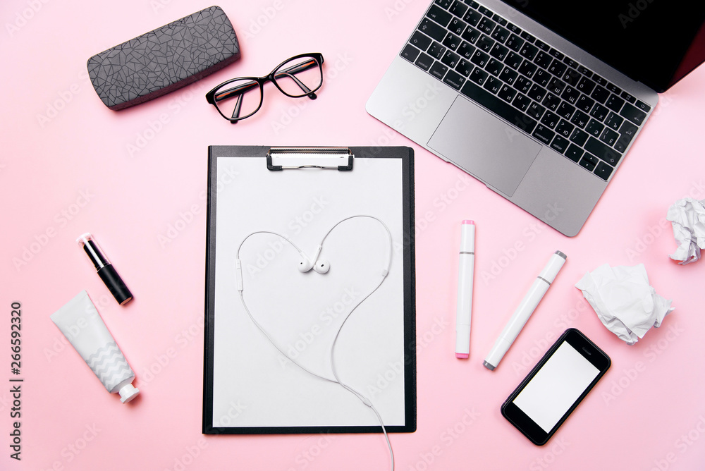 Woman's pink office desk with headphones laid out like a heart. Woman's workplace with laptop, smart phone with blank white screen, a cream, lipstick, eyeglasses and supplies.