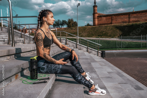 Beautiful girl athlete in tattoos, summer city, resting after fitness workout. Sportswear leggings top. Shaker with water protein skipping rope smartphone headphones. Free space. Listening music.