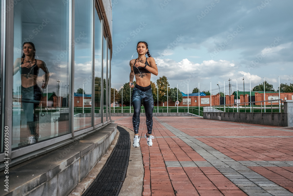 © byswat - Muscular girl runs in the summer in city, on a morning jog, tanned skin, tattoos. Sportswear leggings top. Phone earphones. Fitness concept jogging, lifestyle. Free space for text.