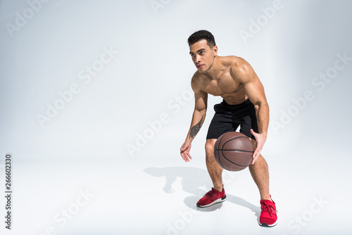 handsome sportive mixed race man in red sneakers playing ball on white