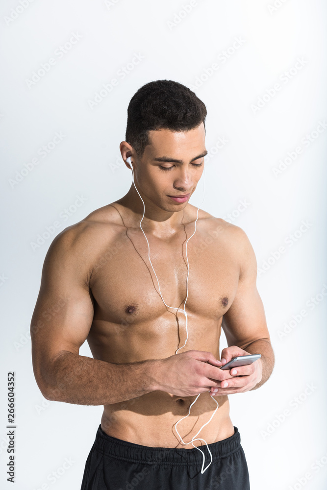 Fototapeta premium handsome sportive mixed race man in earphones holding smartphone and listening music on white