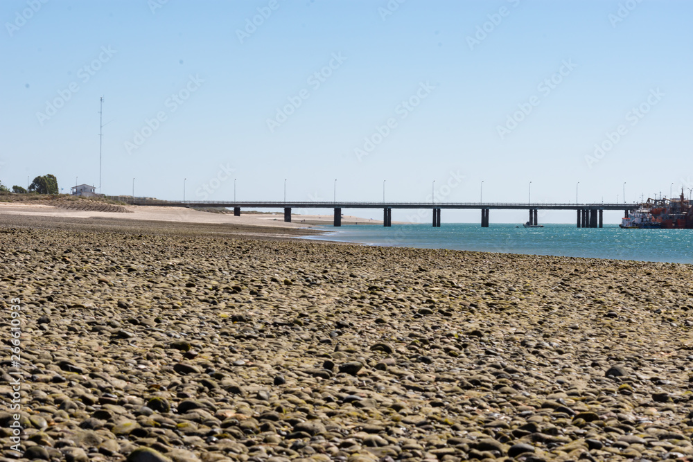 Beach with small stones with pier marking the horizon, sky and blue sea.