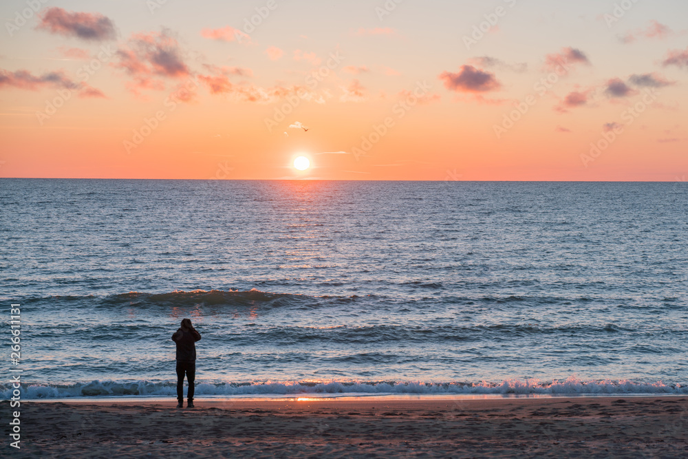 Very beautiful sunset on the Baltic Sea. In the foreground a young man is photographing the illuminated splashing of the waves. The sea is calm.