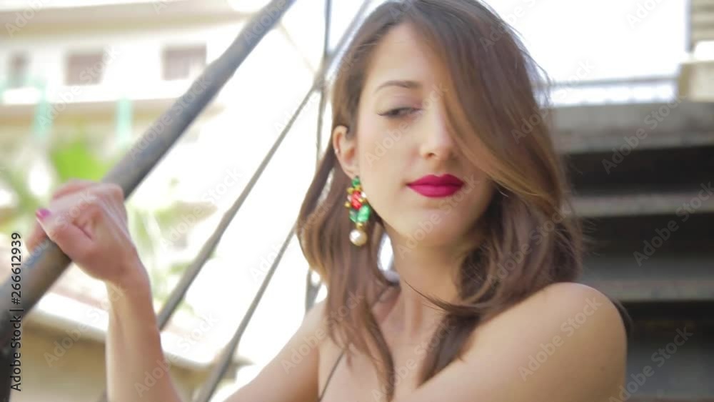 Giovane bella ragazza con capelli lunghi, jeans blu e vestito nero sta posando per foto di moda sulla scala all'esterno di casa vicino al terrazzo.