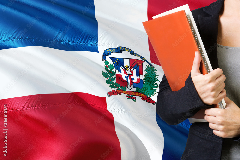 Learning Dominican language concept. Young woman standing with the Dominican Republic flag in the background. Teacher holding books, orange blank book cover.
