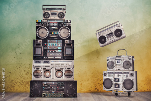 Retro design ghetto blaster boombox cassette tape recorders towers from 1980s on handcart front concrete wall background conceptual composition with flying radio. Vintage old style filtered photo