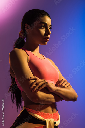 sportive african american girl in pink sports bra posing with crossed hands on purple and blue gradient background