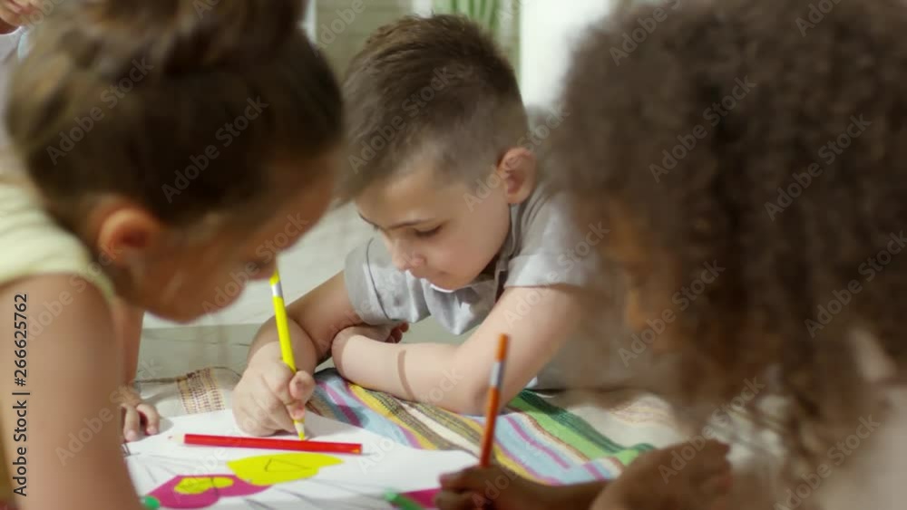 Close up shot of little multiethnic boys and girls lying on the floor and drawing with colored pencils on handmade collage
