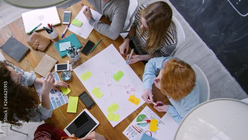 Zoom out top view of team of young colleagues sitting together at office table and drawing business scheme on paper while working together on creative project