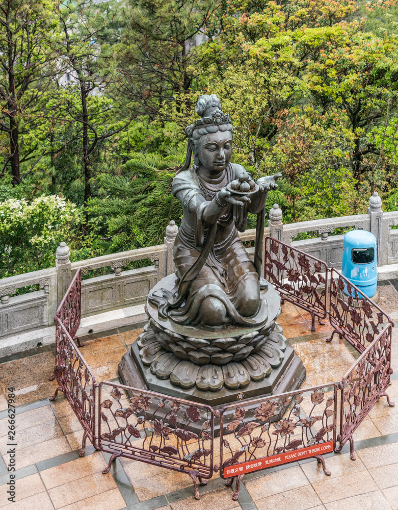 Hong Kong, China - March 7, 2019: Lantau Island. One of the Six Devas offers fruit to Tian Tan Buddha. Bronze statue seen from higher up with green foliage in back.