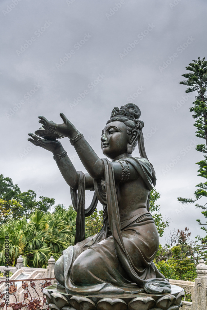 Hong Kong, China - March 7, 2019: Lantau Island. Side Closeup, One of the Six Devas offers fruit to Tian Tan Buddha. Bronze statue seen from front with green foliage and rainy sky in back.