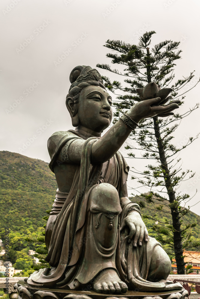 Hong Kong, China - March 7, 2019: Lantau Island. Closeup, One of the Six Devas offers flower to Tian Tan Buddha. Bronze statue seen from front with green foliage and rainy sky in back. Hong Kong, China - March 7, 2019: Lantau Island. Closeup, One of the Six Devas offers flower to Tian Tan Buddha. Bronze statue seen from front with green foliage and rainy sky in back.