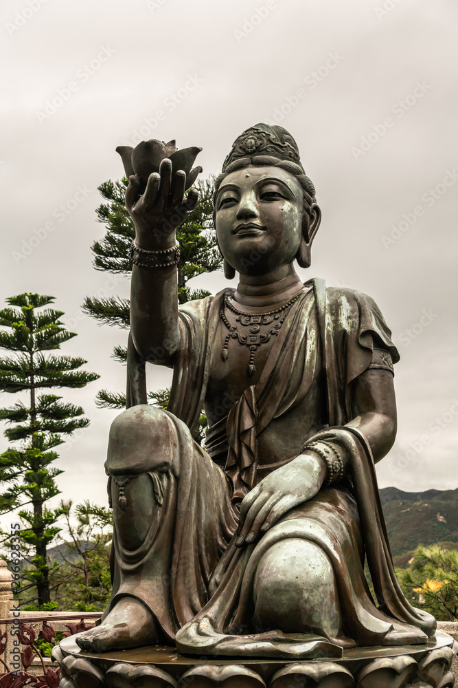 Hong Kong, China - March 7, 2019: Lantau Island. Front Closeup, One of the Six Devas offers flower to Tian Tan Buddha. Bronze statue seen from front with green foliage and rainy sky in back.