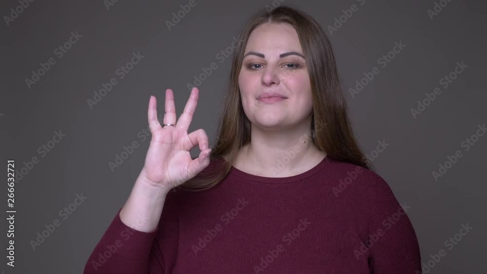 Closeup portarit of young overweight caucasian female gesturing ok handsign looking at camera with background isolated on gray Closeup portarit of young overweight caucasian female gesturing ok handsign looking at camera with background isolated on gray