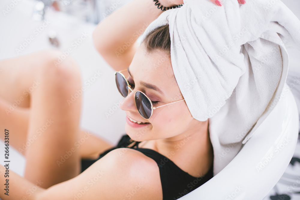 Naklejka premium Young caucasian woman with towel on head and sunglasses getting spa treatment in a beauty salon, inside an interior room. Relaxing in the bathroom in linen. The concept of body care and relaxation