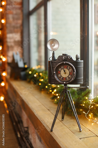 New Year decorations, vintage clock in view of camera. Decorative vintage clock, lanterns with fir-tree garland. Christmas mood. Romantic room in warm colors with a lot of garland lights. Loft style