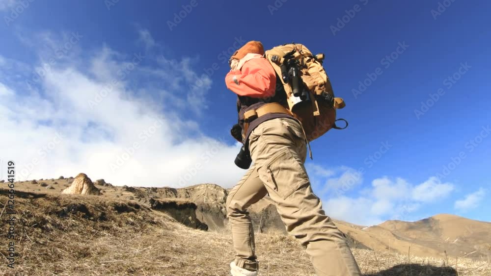 Happy traveler girl photographer in sunglasses with a camera and a backpack goes uphill on a background of snow-capped peaks on a sunny day Happy traveler girl photographer in sunglasses with a camera and a backpack goes uphill on a background of snow-capped peaks on a sunny day