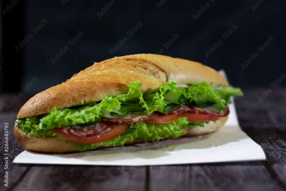 Fresh submarine sandwich with sausage, cheese, bacon, tomatoes, lettuce, cucumbers and onions on a dark wooden background. Mug of beer Fresh submarine sandwich with sausage, cheese, bacon, tomatoes, lettuce, cucumbers and onions on a dark wooden background. Mug of beer