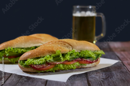 Two fresh submarine sandwiches with sausage, cheese, bacon, tomatoes, lettuce, cucumbers and onions on a dark wooden background. Mug of beer