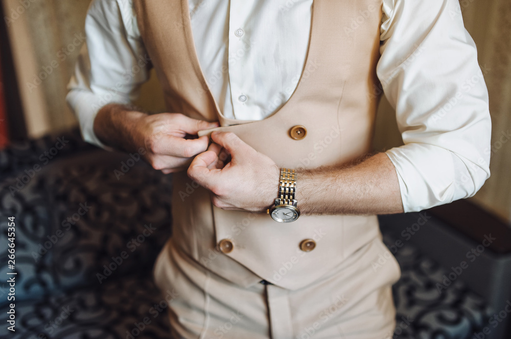 The groom in a beige suit and vest buttons a button. Waiting for the bride. Morning groom. Wedding details. Composition and concept.