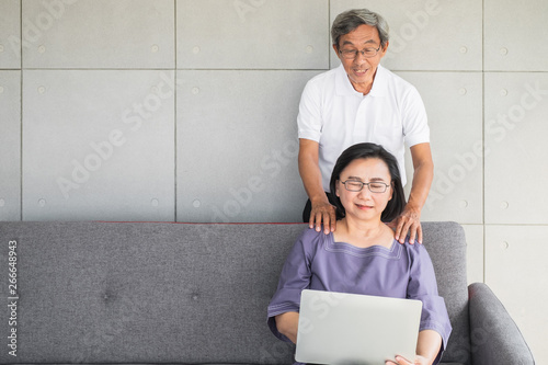 old senior Asian husband giving wife massage while she works on laptop. photo