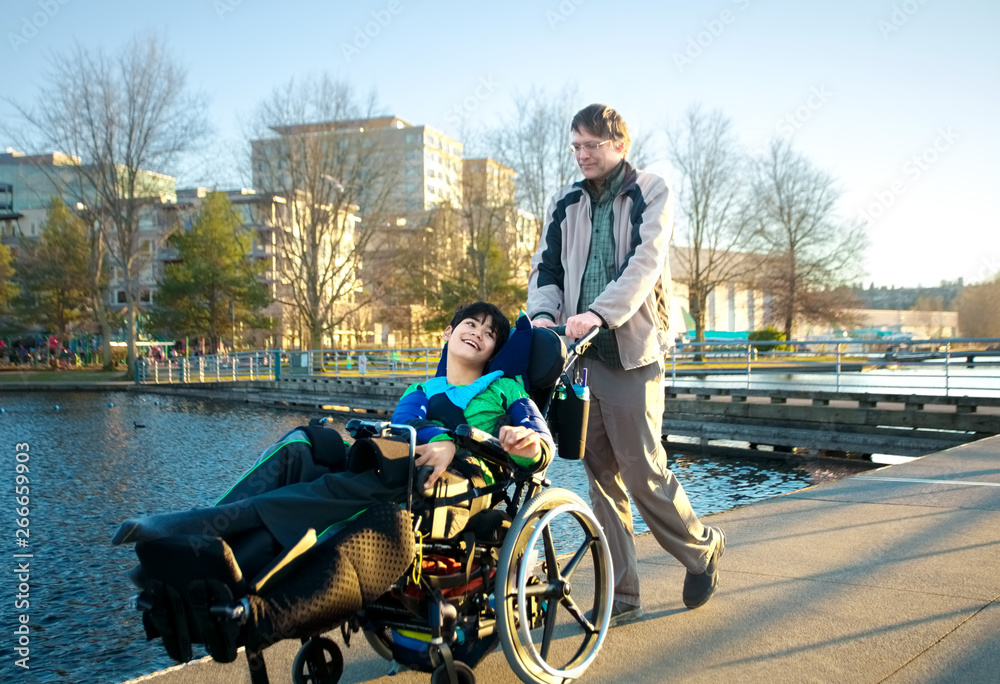 Father walking with disabled son in wheelchair at lake park