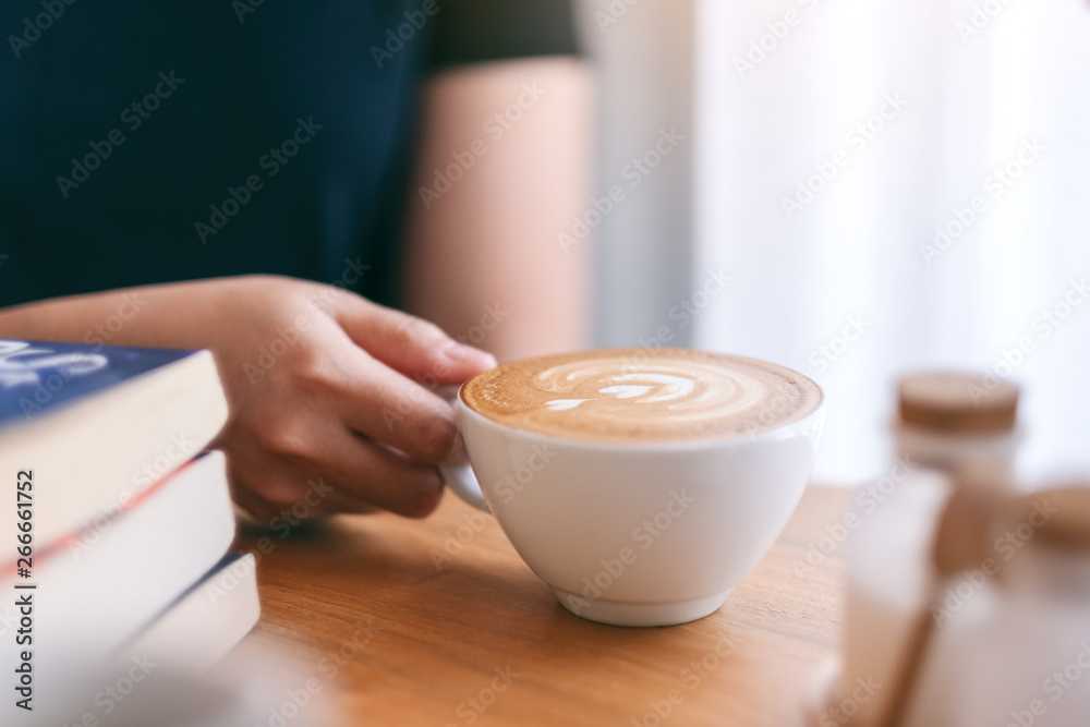 Closeup image of a woman's hand holding a white cup of hot coffee with books on wooden table Closeup image of a woman's hand holding a white cup of hot coffee with books on wooden table